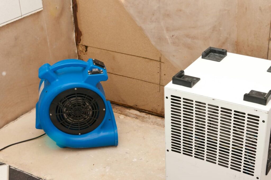Blue air blower fan and white dehumidifier on a floor near a water-damaged wall.