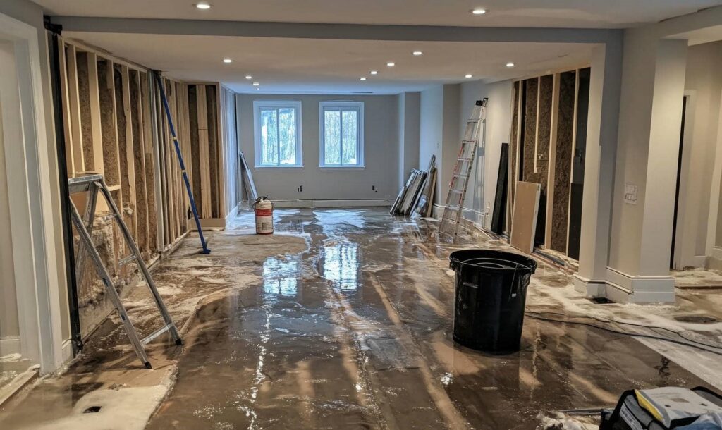 Basement under renovation with exposed walls, wet floor, ladders, and construction materials.